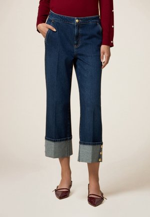 Woman wearing dark blue cropped jeans with wide gray cuffs and gold buttons, paired with burgundy pointed flats and burgundy top.