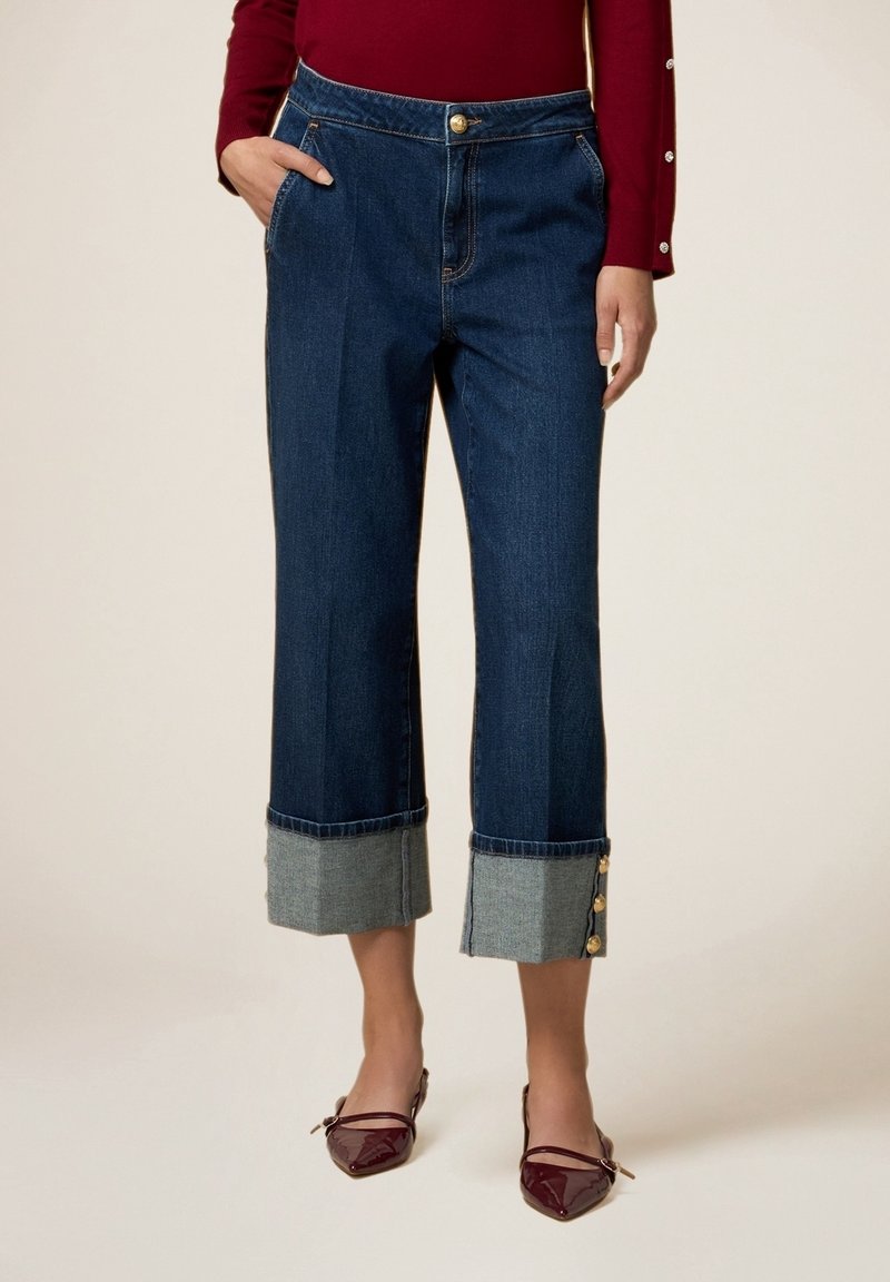 Femme portant un jean bleu foncé court avec de larges revers gris et des boutons dorés, associé à des ballerines pointues bordeaux et un haut bordeaux.