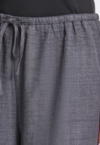 Pantalon gris avec taille élastique et cordon, en tissu texturé, porté avec un haut noir, visible de la taille jusqu’à mi-cuisse.