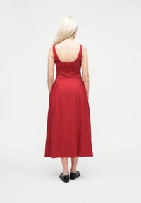 Γυναίκα με μακρι ά ξανθά μαλλιά που φοράει κόκκινο μίντι φόρεμα χωρίς μανίκια και μαύρα χαμηλά παπούτσια, στέκεται με την πλάτη στραμμένη σε απλό λευκό φόντο.