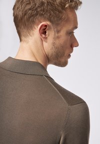 Braunes Strickpolohemd mit Kragen, das über eine gerippte Textur und ein slim-fit Design verfügt, seitlich gezeigt mit kurzem Haar und leichtem Stoppelbart.