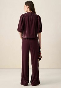 Haut en velours bordeaux avec manches bouffantes et col à volants, associé à un pantalon large assorti. Sac à main noir orné de perles ajoutant une touche de détail.