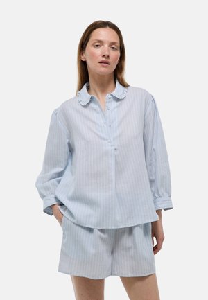 Femme portant un chemisier bleu clair à fines rayures, manches longues avec col à volants et short ample assorti, debout devant un fond uni.