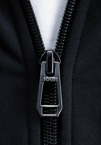 Schwarzer Reißverschluss auf einem dunklen Stoff, mit einem glänzenden metallischen Zipper, auf dem "JOOP!" eingraviert ist, im Kontrast zu einer weißen Innenschicht.