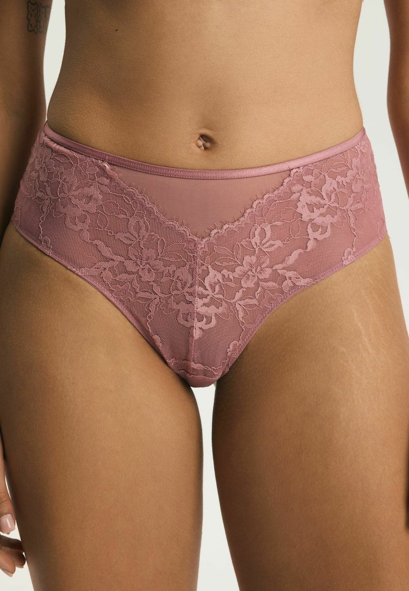 Zbliżenie na środkową część ciała kobiety ubraną w różowe, koronkowe majtki z wysokim stanem i kwiatowymi wzorami na neutralnym tle.