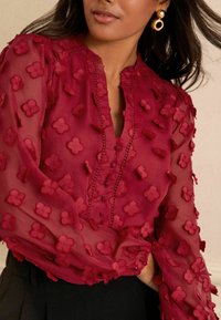 Blusa bordeaux trasparente con applicazioni floreali testurizzate, colletto con bottoni e maniche lunghe. Presenta cuciture sottili e tessuti sovrapposti.