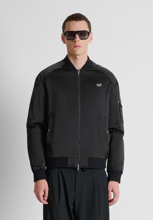 Jonge man die een oversized zwarte bomberjas, zwarte brede broek en grote zwarte zonnebril draagt tegen een effen lichte achtergrond.