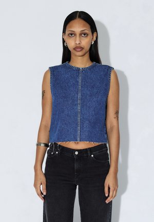 Crop-top uden ærmer i mørkeblå denim med rund halsudskæring, knaplukning foran og opsmøgede kanter. Båret med højtaljede sorte jeans.