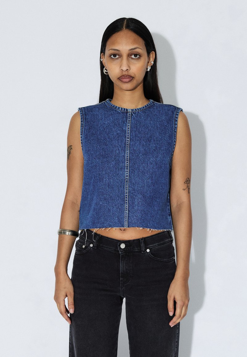 Crop-top uden ærmer i mørkeblå denim med rund halsudskæring, knaplukning foran og opsmøgede kanter. Båret med højtaljede sorte jeans.