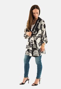 Kimono en blanco y negro con estampado de hojas de palma, ceñido a la cintura. La modelo lo lleva con una blusa blanca y jeans ajustados azules, acompañados de tacones negros.