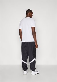 T-shirt branca e calças de treino pretas com uma faixa branca. As calças de treino têm um painel em rede e um corte relaxado. O modelo usa tênis brancos de desporto.
