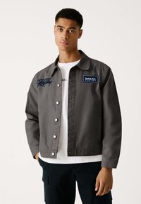 Graue Canvas-Jacke mit Druckknöpfen, Details des Red Bull Racing-Logos und einem Kragendesign. Über einem weißen T-Shirt getragen.