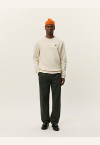 Kremowa bluza z małym haftowanym wzorem, zestawiona z ciemnozielonymi spodniami i jasnopomarańczową wełnianą czapką. Czarne buty dopełniają outfit.
