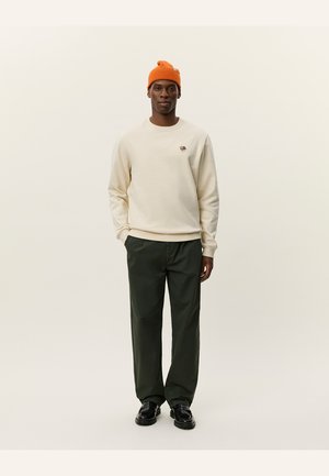 Kremfarget sweatshirt med et lite brodert design, parret med mørkegrønne bukser og en lys oransje strikket lue. Svarte sko fullfører antrekket.