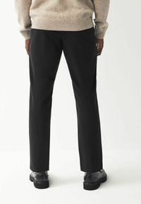 Pantalon noir à coupe droite, fabriqué en tissu lisse, associé à des chaussures noires. Bas de pull côtelé visible à la taille.