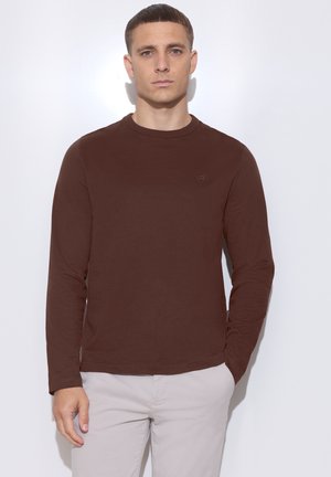 Joven con el pelo corto, vestido con una camisa marrón de manga larga y pantalones beige claros, de pie frente a un fondo blanco con expresión neutral.
