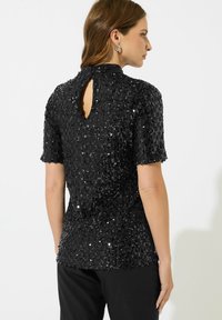 Schwarzes Paillettenoberteil mit kurzen Ärmeln, strukturiertem Design, hohem Ausschnitt und einem Schlüssellochausschnitt auf der Rückseite, kombiniert mit schwarzen Hosen.