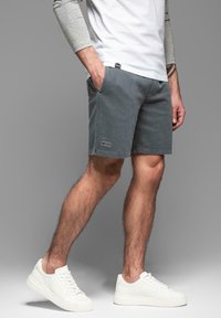 Pantalones cortos de algodón grises con dos bolsillos laterales y un pequeño parche de logo. Combinados con zapatillas blancas, que presentan una textura suave y un diseño clásico.