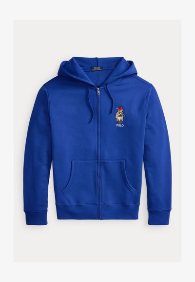 Modrá mikina na zip vyrobená z měkké látky, s klokáním kapsou a vyšitým medvědem s červenou čepicí na levé straně hrudi.