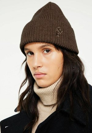 Persona joven con cabello oscuro y largo, que lleva un gorro de punto marrón, un jersey de cuello alto beige y un abrigo negro sobre un fondo liso.