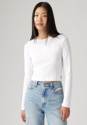 Valkoinen pitkähihainen cropped-paita, jossa on Levi's-logo rinnassa, yhdistettynä vaaleansinisiin korkeavyötäröisiin kulutettuihin farkkuihin.