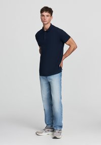Giovane uomo in piedi con le mani dietro la schiena, che indossa una polo blu navy, jeans azzurri e sneakers grigie su uno sfondo neutro.