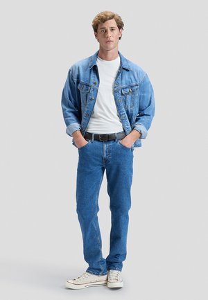 Ung mand står med hænderne i lommerne iført en lyseblå denimjakke, hvid t-shirt, blå jeans, sort bælte og hvide sneakers.