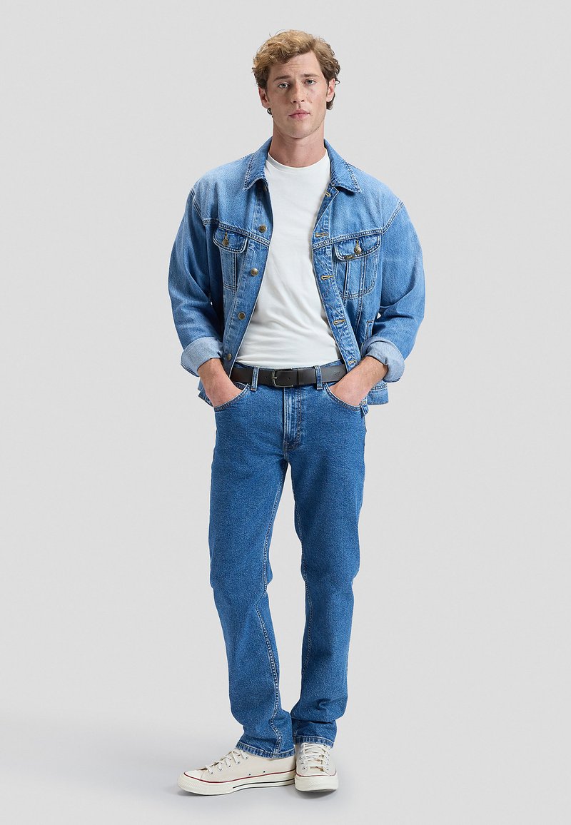 Junger Mann steht mit den Händen in den Taschen, trägt eine hellblaue Jeansjacke, ein weißes T-Shirt, blaue Jeans, schwarzen Gürtel und weiße Sneakers.