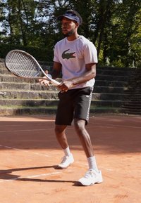 Tenisist v sivi Lacoste majici z logotipom krokodila, črnih kratkih hlačah in belih športnih copatih, drži teniško loparico na školjčnem igrišču.