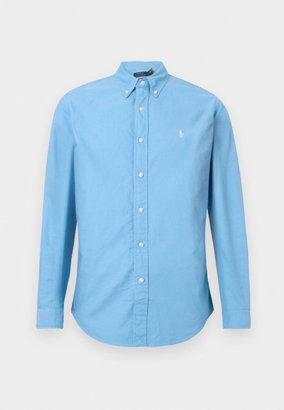 Camisa de botão de manga longa azul-claro. Tecido de algodão, colarinho tipo botão, cinco botões na frente, com um pequeno logótipo amarelo no lado esquerdo do peito.