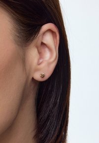 Złota kolczyk w kształcie róży, z gładkim wykończeniem. Na jasnym tle, które podkreśla kształt i detale kolczyka.