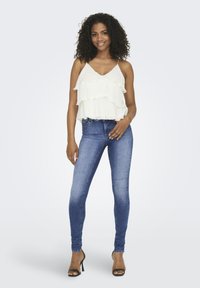 Haut blanc à volants sans manches avec des fines bretelles, associé à un jean skinny bleu et des sandales à talons hauts noires. Texture lisse dans l'ensemble.