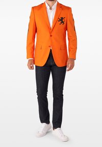 Pomarańczowa marynarka z czarnym emblematem lwa na piersi, z zapięciem na jeden guzik, z klapami, noszona na białej koszuli, w połączeniu z ciemnymi dżinsami i białymi trampkami.
