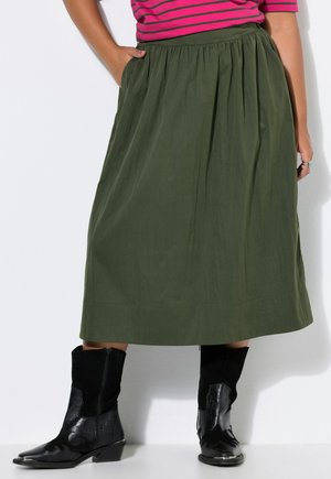 Mujer con falda midi verde oliva, botas negras hasta la mitad de la pantorrilla y una camiseta de rayas rosa y negra con una mano en el bolsillo.