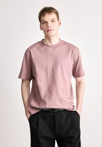 Světle růžové tričko s krátkým rukávem vyrobené z bavlny, s kulatým výstřihem a malým vyšívaným logem "HUGO" na hrudi.