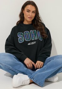 Sudadera negra con "SOHO est. 1978" en letras audaces y coloridas. Combinada con jeans holgados de color azul claro y zapatillas blancas. Textura de tela suave.