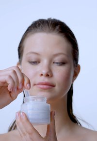 Un pot en verre blanc de masque facial avec du texte bleu et une spatule transparente, tenu par une personne aux longs cheveux mouillés, sur un fond clair.