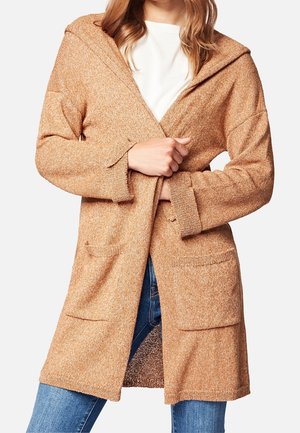 Vrouw draagt een beige gebreid vest met open voorkant en zakken, over een wit shirt en blauwe jeans, met handen die de voorste kanten vasthouden.