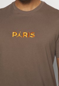 Hnědé bavlněné tričko s vyšitým oranžovým logem "PARIS" a grafikou Jumpman umístěnou centrálně pod textem. Krátké rukávy a kulatý výstřih.
