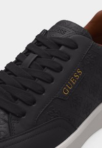 Gros plan sur une basket noire Guess avec des motifs embossés, des lacets noirs et le logo "GUESS" doré sur le côté, au-dessus d'une semelle blanche.