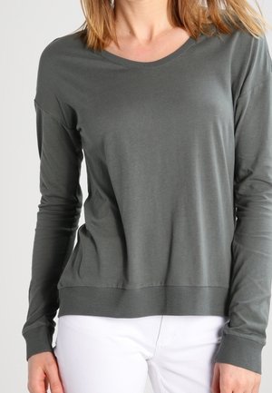 Mujer con una camisa gris oscuro lisa de manga larga y cuello redondo, y pantalones blancos, de pie frente a un fondo neutro.
