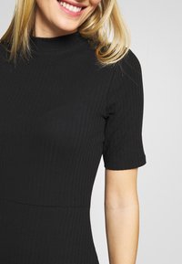 Robe noire à côtes avec un col haut et des manches courtes. Présente un design ajusté avec une texture lisse. Le bras du mannequin est partiellement visible.