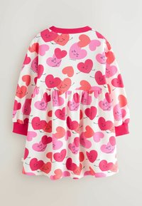 Pitkähihainen puuvillamekko, jossa on valkoinen tausta ja erilaisia vaaleanpunaisia sydäntemoja hymyilevin kasvoin. Raskas punainen reunus kaula-aukkossa.