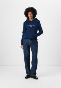 Femeie stând pe fundal alb, purtând hanorac cu glugă bleumarin cu logo, blugi largi albaștri și mocasini negri, cu o mână în buzunar.