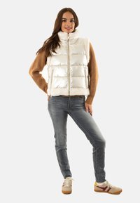 Gilet bianco e gonfio con finitura lucida e morbido bordo in pelliccia; abbinato a un maglione marrone, jeans grigi e sneaker beige con dettagli rossi.