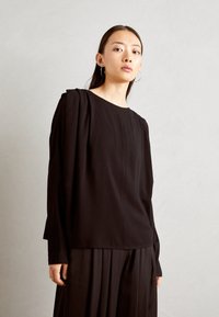 Top nero a maniche lunghe con scollatura rotonda e dettaglio delle spalle plissettato, realizzato in tessuto morbido; abbinato a pantaloni neri plissettati.