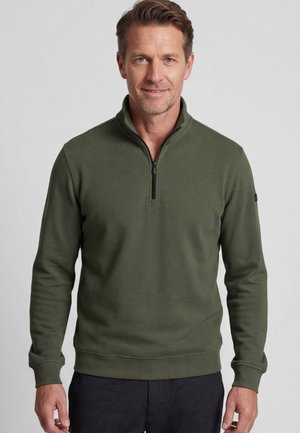 Man wearing olive green quarter-zip pullover sweater and dark pants, standing against plain light gray background.