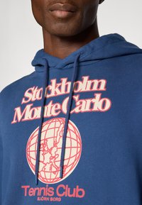 Közelkép egy olyan személyről, aki kék kapucnis pulóvert visel, piros és fehér "Stockholm Monte Carlo Tennis Club" felirattal, valamint egy földgömb grafikával.