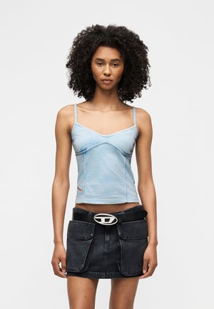 Žena s kučeravými vlasmi, ktorá má na sebe svetlomodrý camisole top a čiernu rifľovú mini sukňu s veľkými vreckami vpredu a čiernym opaskom.