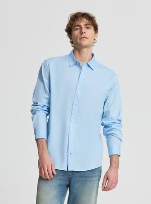 Giovane uomo con capelli castani spettinati, indossa una camicia azzurra con bottoni e jeans blu sbiaditi, in piedi davanti a uno sfondo semplice.